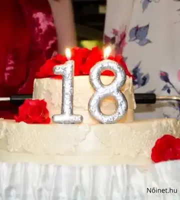 Ezen a képen egy fehér fondant bevonatú torta látható, amelyet ezüst színű "18" számú gyertya díszít. A tortát piros rózsák díszítik, és a háttérben ünneplő emberek láthatók, elegáns ruhában.